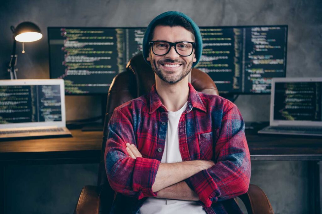 Photo of happy coder dressed in a hat, glasses, and flannel shirt, smiling, arms folded, indoors sitting at his workstation